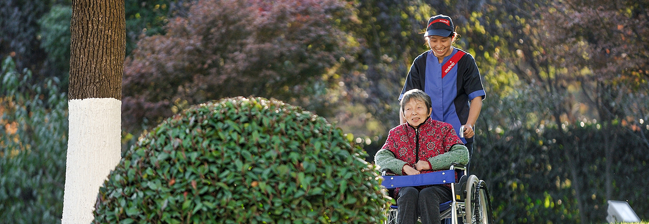 Sodexo-Mitarbeiter schiebt eine ältere Frau im Rollstuhl durch einen Garten.